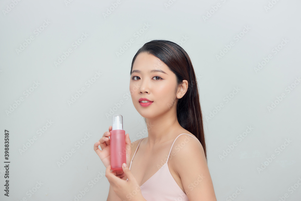 Young pretty stylish woman over white wall background holding bottle with cosmetics water. Young pretty stylish woman over white wall background holding bottle with cosmetics water.