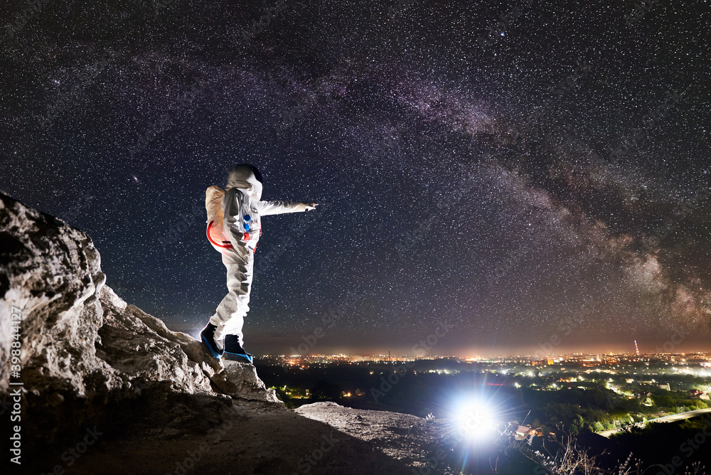 Space traveler pointing finger at night city while standing on rocky hill under majestic sky with stars. Mission specialist astronaut wearing white space suit. Concept of human space exploration.