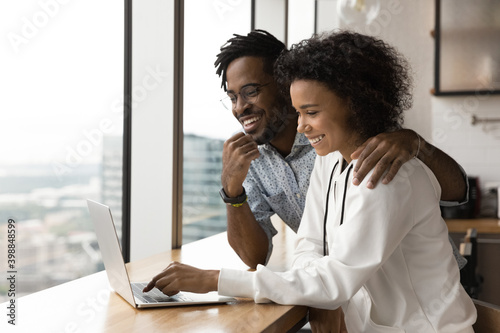 Fotografia Happy African American couple sit at table at home look at laptop screen browse internet together