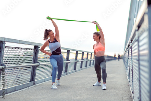 Happy fitness female friends in sportswear stretch arms together with rubber band