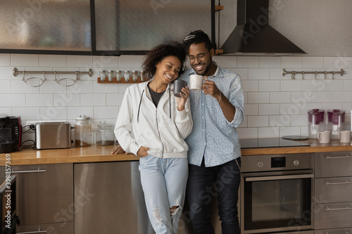 Overjoyed millennial biracial family man and woman relax drink coffee together in new renovated home kitchen. Happy young African American couple renters enjoy moving relocation to own house.