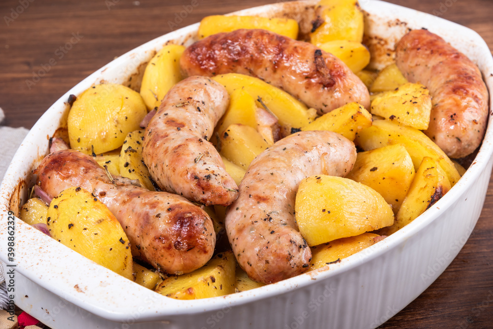 Baked golden pieces of potatoes with turkey sausages in a white ceramic baking dish on a wooden table, close-up Baked golden pieces of potatoes with turkey sausages in a white ceramic baking dish on a wooden table, close-up