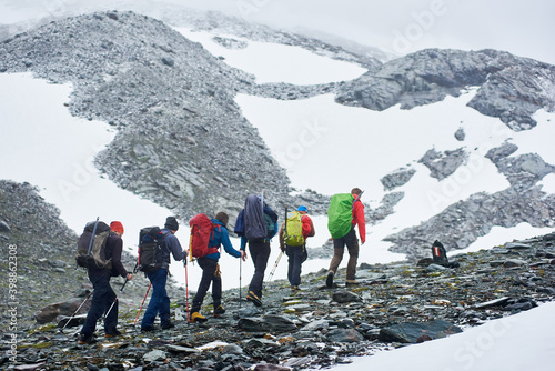 Back view of male hikers with backpacks walking on rocky path in winter mountains. Group of mountaineers with trekking sticks climbing the rocky hill. Concept of travelling, hiking and mountaineering.
