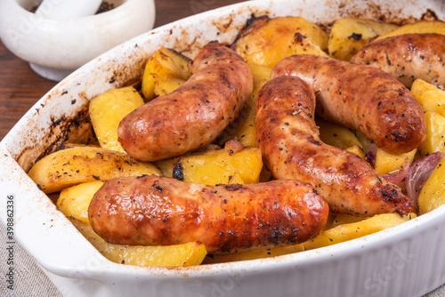 Baked golden pieces of potatoes with turkey sausages in a white ceramic baking dish on a wooden table, close-up
