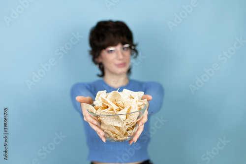 Young beautiful woman holding nachos potato chips over isolated blue background smiling and showing the bowl at the camera