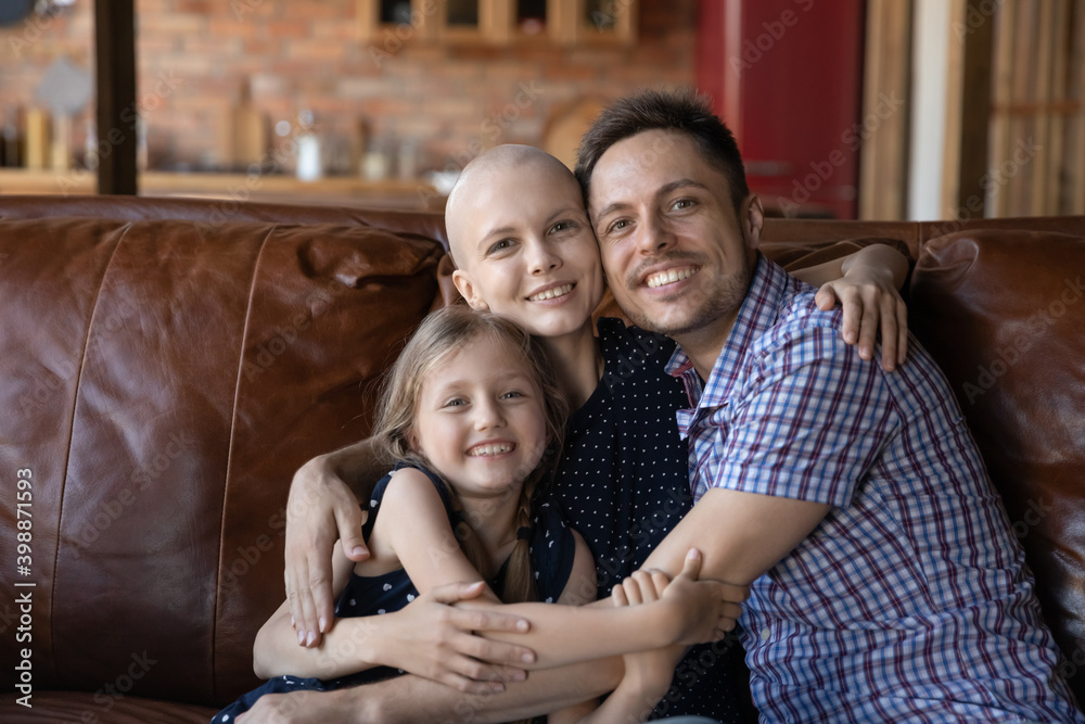 Fototapeta premium Portrait happy sick mother cancer patient, father and little daughter cuddling, looking at camera, smiling unhealthy woman and man with child posing for photo together, showing support and care