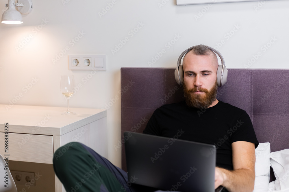 Caucasian bearded man in headphones in bedroom on bed working on laptop from home, typing, thinking. Caucasian bearded man in headphones in bedroom on bed working on laptop from home, typing, thinking.
