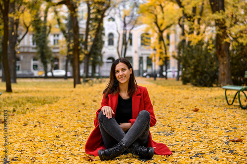 Woman in red coat sitting in park and smiling to photographer. Autumn season.