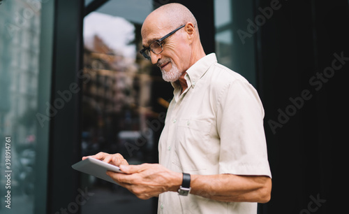 Mature male blogger 60 years old reading network publication in social media using touch pad at urban setting, Caucasian man in eyeglasses connecting to 4g wireless for making booking and banking