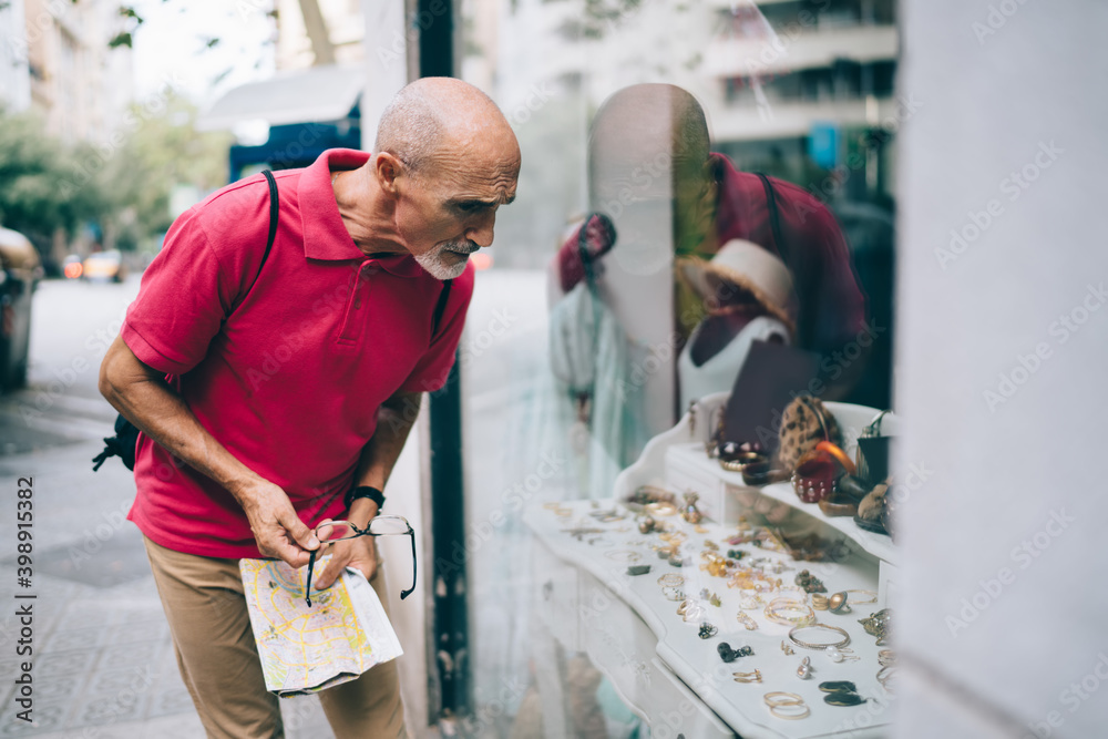 Mature tourist with travel map in hands standing near store showcase choosing handmade jewelry for buying present, adult Caucasian man searching retail in giftshop during solo vacations on retirement Mature tourist with travel map in hands standing near store showcase choosing handmade jewelry for buying present, adult Caucasian man searching retail in giftshop during solo vacations on retirement