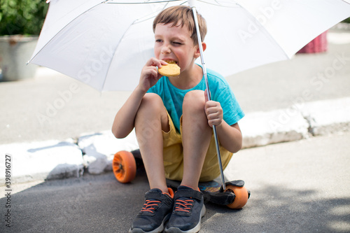 Cheerful boy in a blue t-shirt sits on a skateboard under a white umbrella