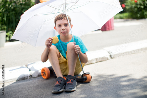 Cheerful boy in a blue t-shirt sits on a skateboard under a white umbrella
