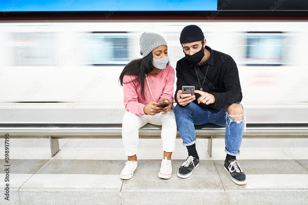 A young interracial couple of lovers with masks and wool hats sits on the subway platform looking at something interesting smart phone. The movement of the subway caravan.