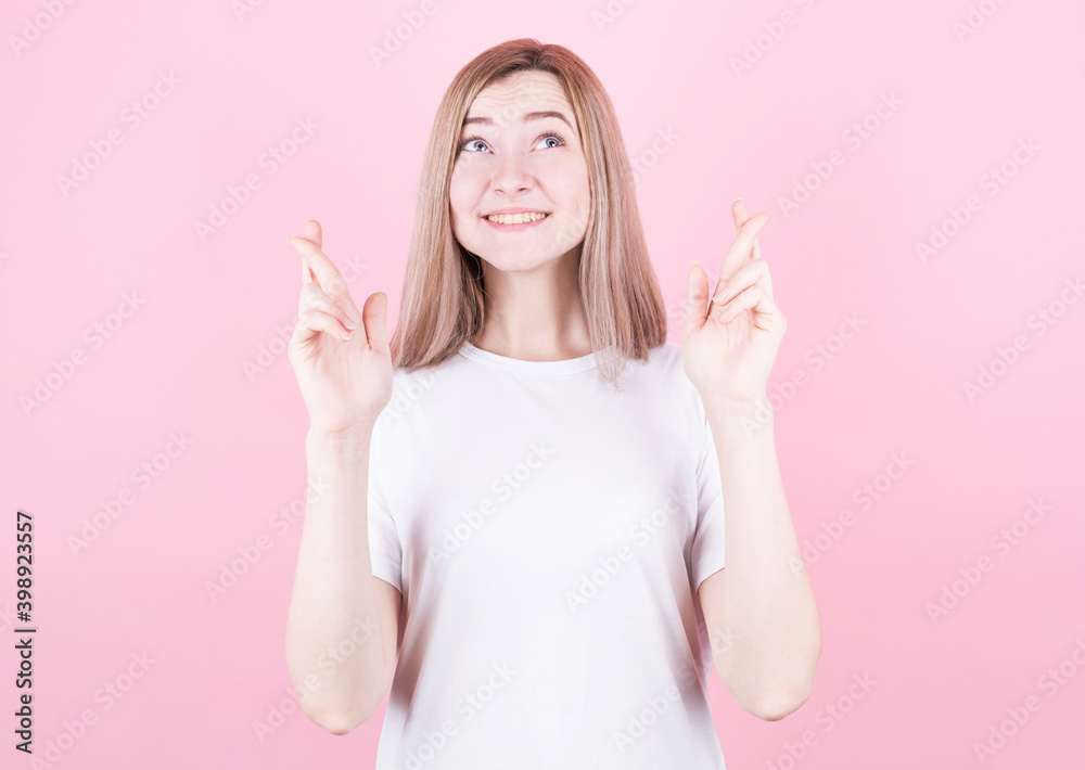 Closeup of happy lovely young woman wears t shirt keeps fingers crossed and looks up, isolated over pink background Closeup of happy lovely young woman wears t shirt keeps fingers crossed and looks up, isolated over pink background