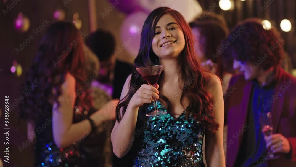 Dancing lady in a sequin dress at stuff party in front of the camera feeling relaxed and happy holding glass of drink and smiling large Dancing lady in a sequin dress at stuff party in front of the camera feeling relaxed and happy holding glass of drink and smiling large