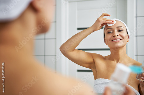 Smiling woman applying micellar water and cleaning her face. Reflection in the mirror