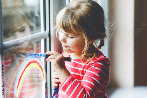 Adoralbe little toddler girl with rainbow painted with colorful window color during pandemic coronavirus quarantine. Child painting rainbows and hearts around the world with words Let's all be well.