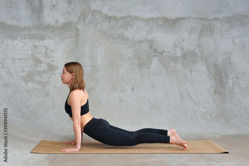 Beautiful woman stretching in Cobra pose, doing Bhujangasana exercise, practicing yoga, attractive sporty girl wearing black sportswear working out at home or in yoga studio.