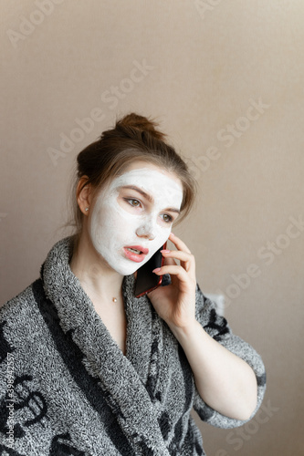 a young girl takes care of her skin and talks on a mobile phone, in a gray terry robe and a clay mask on her face