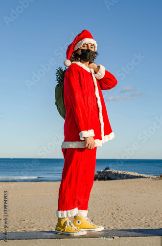 A vertical shot of Young Santa Holding Green Bag With Beach Background at Christmas.