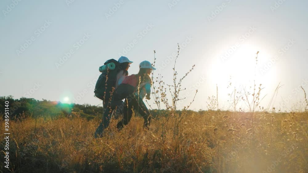 Mother and daughter with backpacks running through the meadow at sunset. Family tourism concept. Mother and daughter with backpacks running through the meadow at sunset. Family tourism concept.