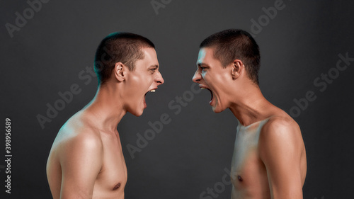 Two young half naked angry caucasian twin brothers arguing, shouting while standing face to face isolated over dark grey background