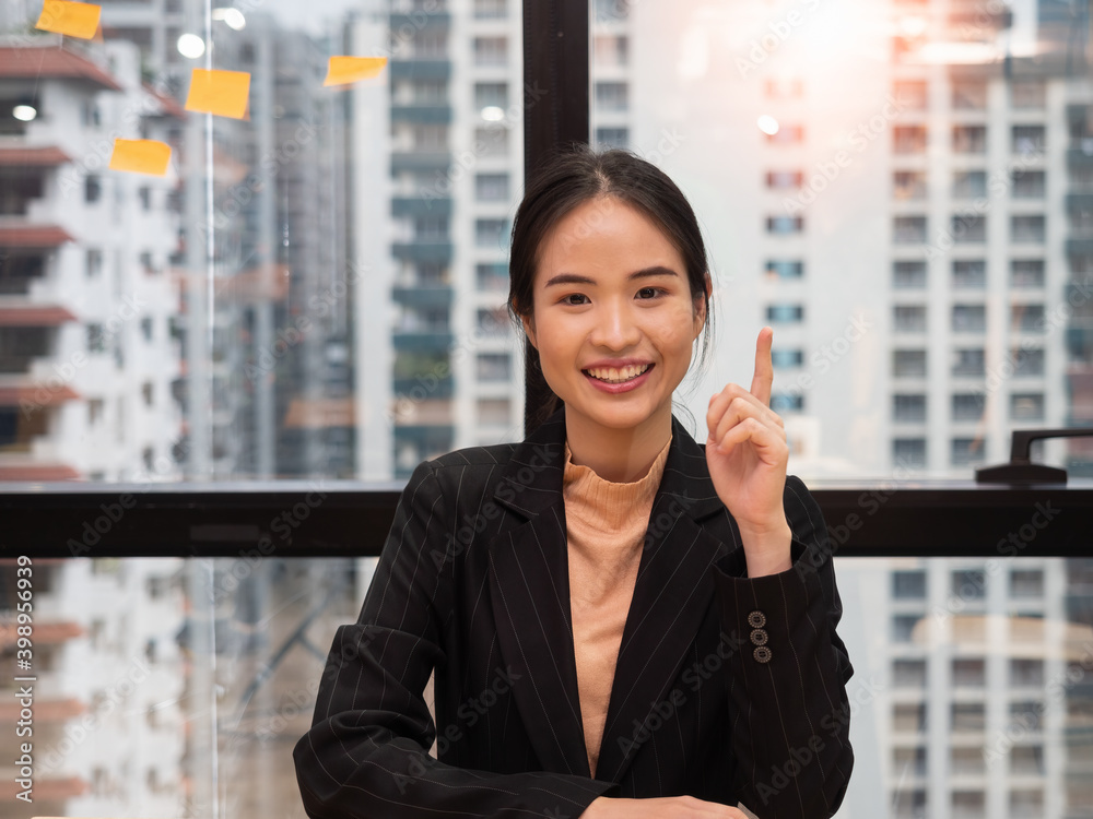Portrait of confident smiling happy pretty young Asian woman wearing a black suit sitting at the desk in the office. Portrait of confident smiling happy pretty young Asian woman wearing a black suit sitting at the desk in the office.