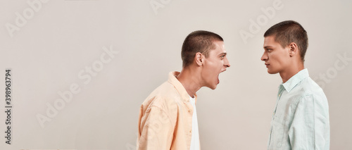 Portrait of two young caucasian men, twin brothers arguing, shouting while standing face to face isolated over beige background, panoramic banner