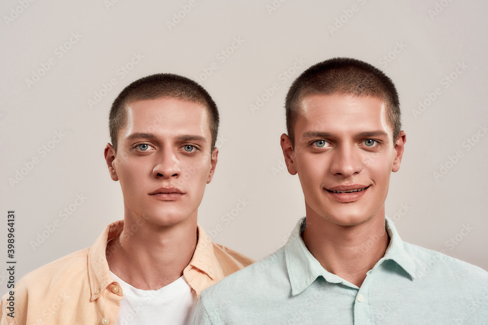 Sadness and happiness. Close up portrait of two young caucasian twin brothers looking at camera with different face expressions while standing isolated over beige background Sadness and happiness. Close up portrait of two young caucasian twin brothers looking at camera with different face expressions while standing isolated over beige background