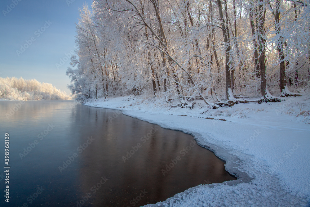 Winter day on the river, partially covered with ice. On the shore there are trees in hoarfrost.