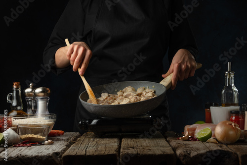 Professional chef stir frying chicken fillet in the pan. Backstage of cooking traditional Indian chicken curry on dark blue background. Frozen motion. Concept of cooking process.