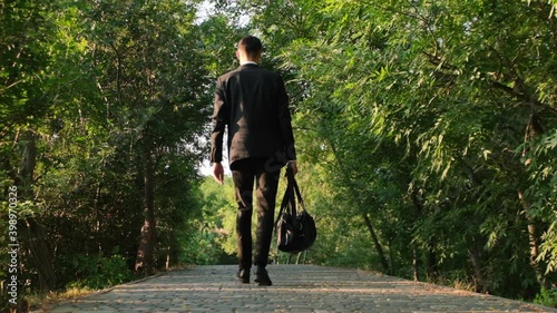 A man in a business suit walks along the road in the park with a travel bag in his hand. Goes with his back to the video camera, shooting 120 fps.