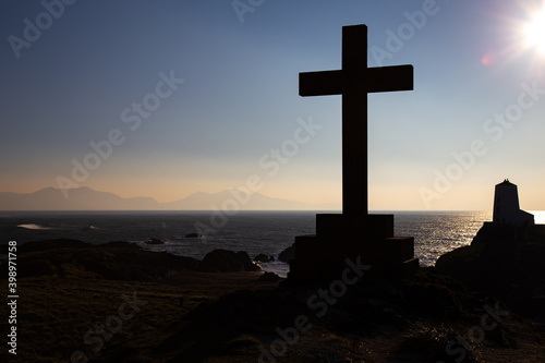 Cross of Dwywen and Llanddwyn Lighthouse, Llanddwyn Island, Anglesey, Wales. Near Newborough Sands. Silhouette against a sunset sky with Snowdonia mountain rage in the background.