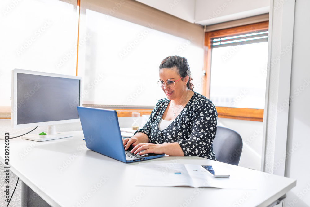 woman in office with blue laptop in white office with lots of light and large veins working at desk woman in office with blue laptop in white office with lots of light and large veins working at desk