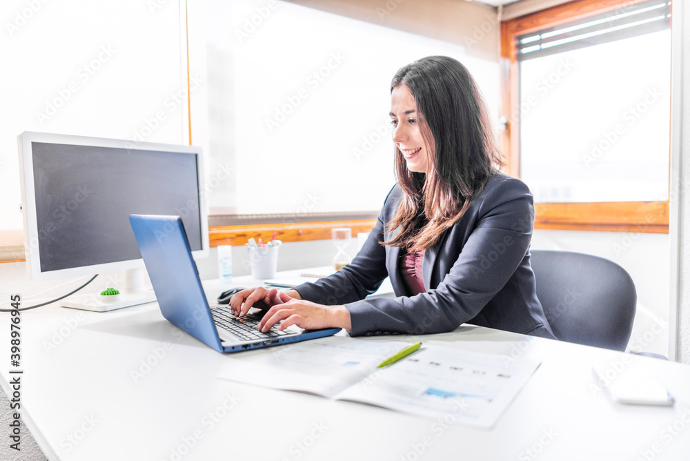 woman in office with blue laptop in white office with lots of light and large veins working at desk woman in office with blue laptop in white office with lots of light and large veins working at desk