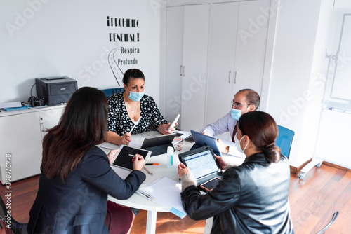 business meeting 4 people with laptops ipad tablet folios pens and folios in bright office with large windows three women and a man happy smiling covid mask