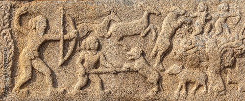 Hampi, Karnataka, India - November 4, 2013: Mural sculpture on brown stone at Royal Enclosure. Closeup of hunting scene with people and deer. Bow and spear as weapons.