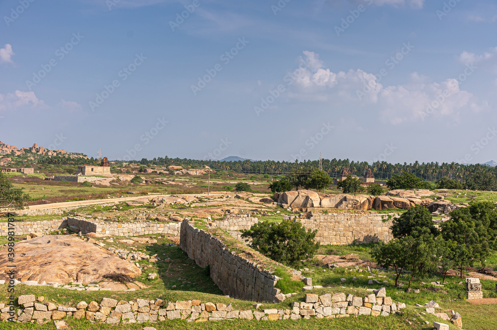 Hampi, Karnataka, India - November 4, 2013: Wide shot over fundaments and parts of wall is what mostly stands in Royal Enclosure. Blue cloudscape and green foliage dispersed among beige-brown stones.