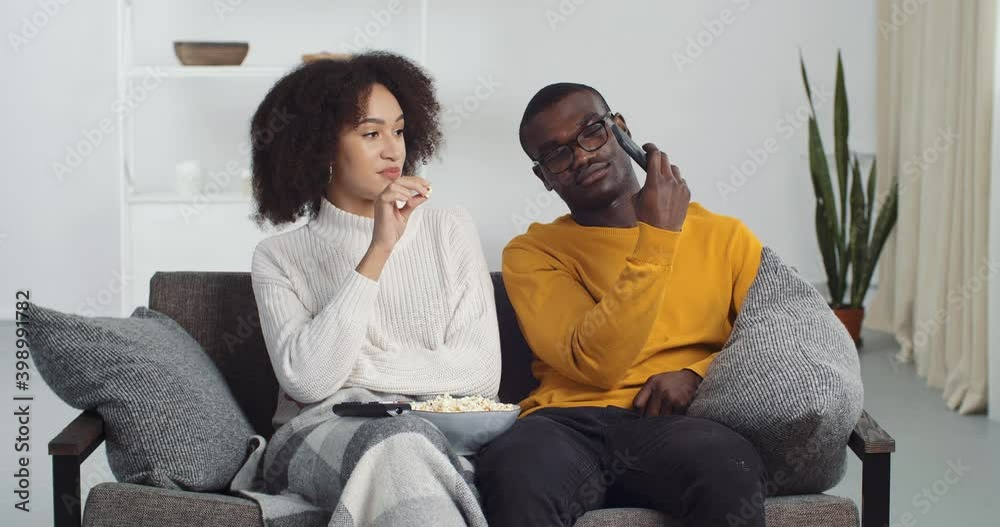 Couple of teenagers afro american young newlyweds husband and wife sitting at home on couch listening to music eating popcorn enjoying song watching music videos on mobile smart phone online dancing Couple of teenagers afro american young newlyweds husband and wife sitting at home on couch listening to music eating popcorn enjoying song watching music videos on mobile smart phone online dancing