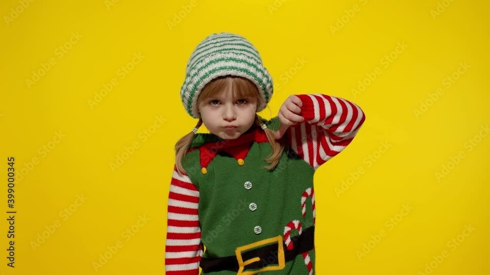 Displesed blonde kid teen girl in Christmas elf Santa helper costume doesnt agree with ideas how to celebrate New Year keeps thumb down shows dislike gesture isolated on yellow background. Slow motion Displesed blonde kid teen girl in Christmas elf Santa helper costume doesnt agree with ideas how to celebrate New Year keeps thumb down shows dislike gesture isolated on yellow background. Slow motion