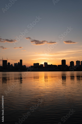 夜明け前、大阪淀川河川敷から梅田の高層ビルをのぞむ。オレンジ色に染まった空に摩天楼がシルエットになり水面に映る