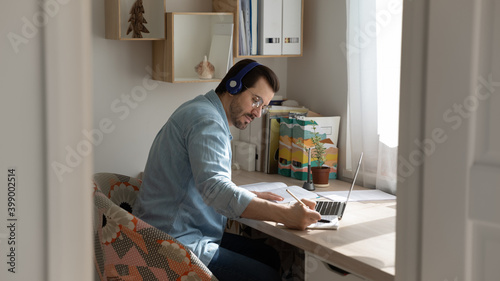 Focused man wearing glasses and headphones studying online, writing notes, using laptop, sitting at work desk, student listening to lecture, learning language, businessman watching webinar