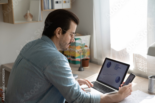 Fototapeta Naklejka Na Ścianę i Meble -  Close up serious businessman wearing glasses working with project statistics at home, preparing presentation with graphs and diagrams, using laptop and smartphone, accountant analyzing financial data
