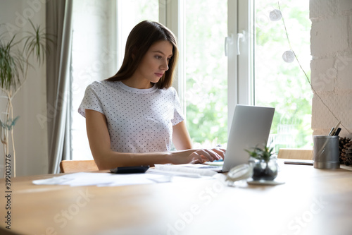Focused young woman checking financial documents, using laptop, browsing online banking services at home, sitting at desk with papers and calculator, domestic bills or expenses, planning budget