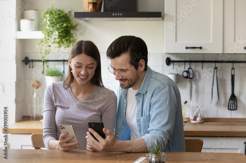 Happy young couple using smartphones, sitting at wooden table in modern kitchen, smiling wife and husband showing funny videos or photos in social network, spending leisure time with gadgets
