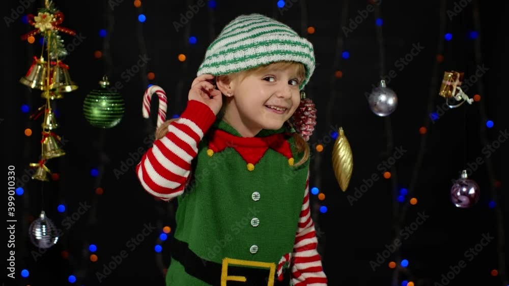 Kid girl in Christmas elf Santa Claus helper costume keeps hand near ear tries to overhear what present gifts parents going to give her. Child interested in someones conversation. New Year holidays Kid girl in Christmas elf Santa Claus helper costume keeps hand near ear tries to overhear what present gifts parents going to give her. Child interested in someones conversation. New Year holidays