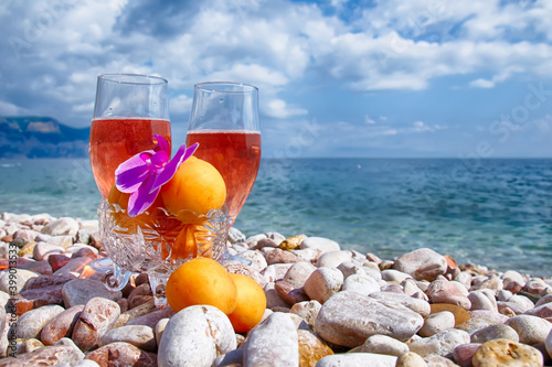 Two glasses with a cocktail and tropical flowers, a vase with fresh fruit stand side by side on a large white sea pebble against the sea background. Summer.
