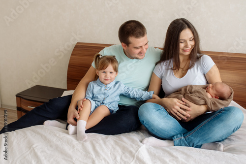 Young beautiful caucasian family with a three-year-old daughter and a newborn toddler sits embracing on the bed in the bedroom at home 