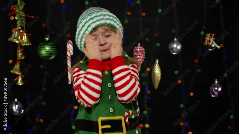 Teen kid girl in Christmas elf Santa Claus helper costume playing hide-and-seek isolated on black background with garland. Child cover face with hands, fooling around. New Year holidays celebration Teen kid girl in Christmas elf Santa Claus helper costume playing hide-and-seek isolated on black background with garland. Child cover face with hands, fooling around. New Year holidays celebration