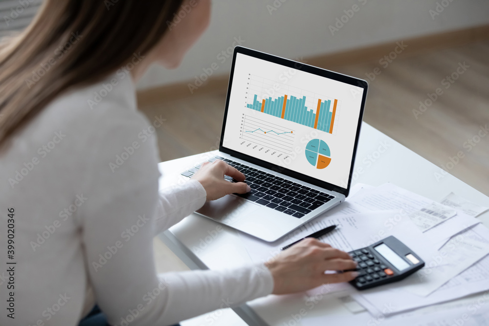 Close up rear view young woman using laptop and calculator, working on project, checking financial documents, sitting at table at home, looking at computer screen with diagrams, calculating bills